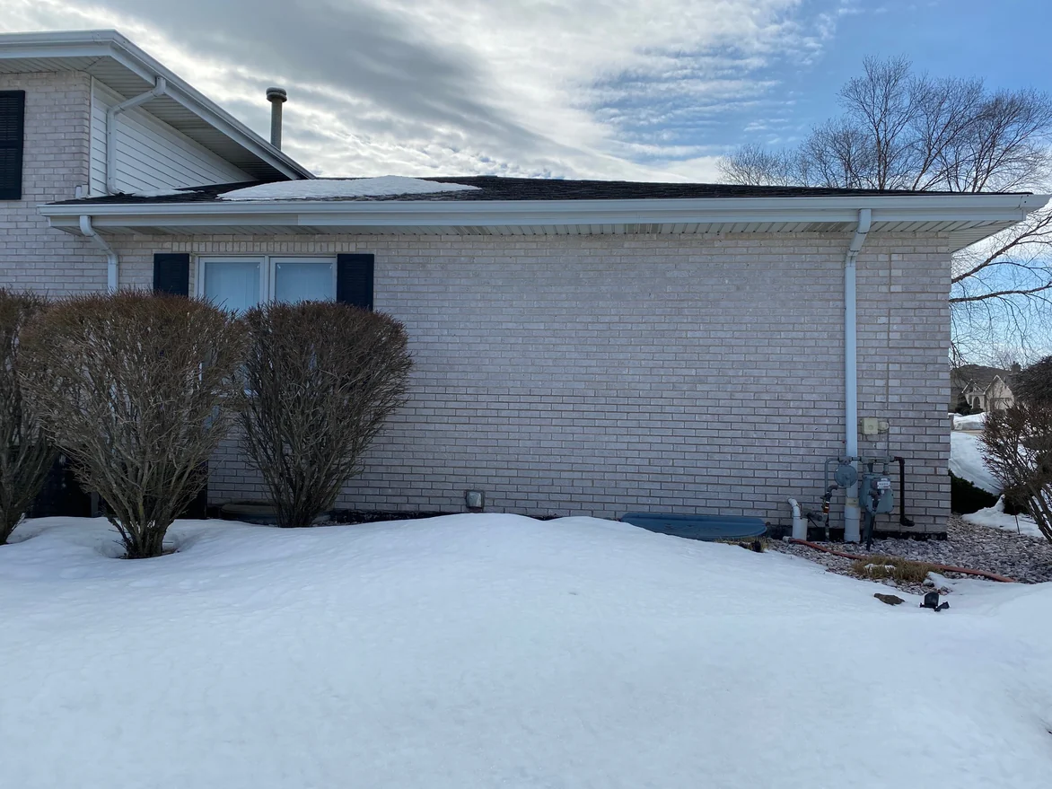 The side wall of the single-story wing in 2021, pale brick running from a shuttered window at the left to the corner pier at the right where the gas meter stands, with bare shrubs and deep snow in front