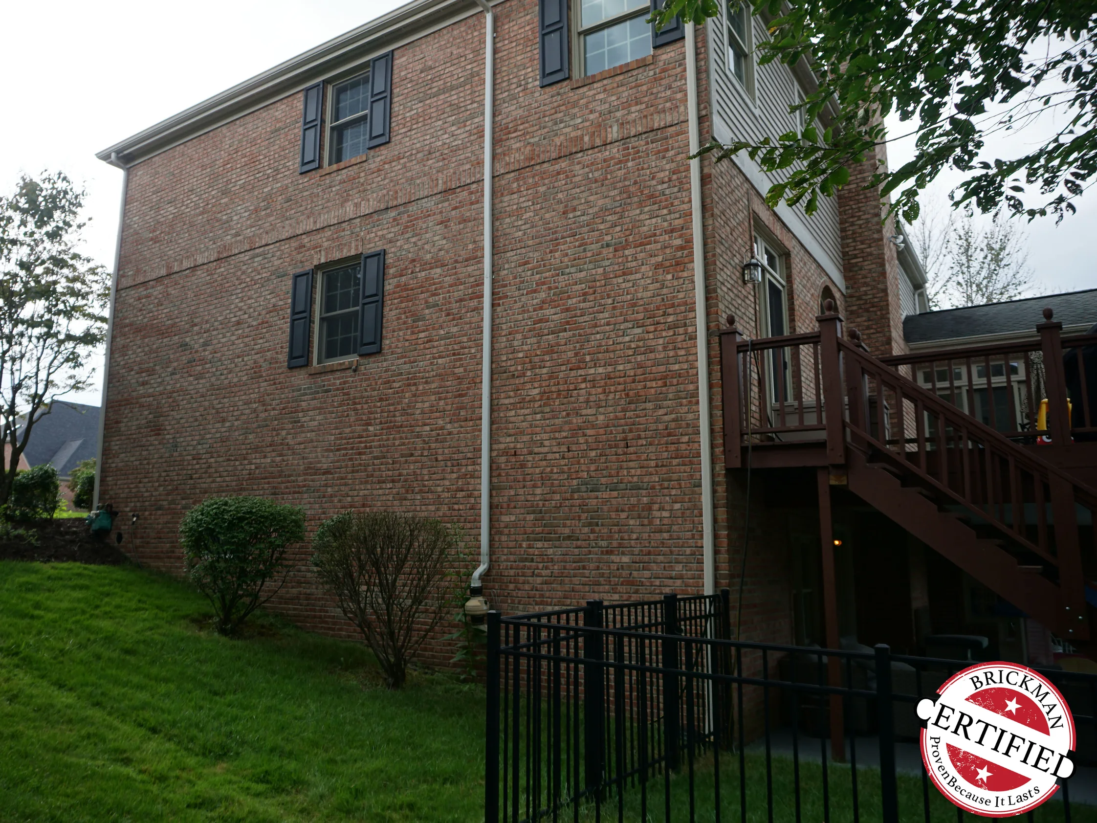 The side elevation in 2019, brick running from the eaves down the slope past a raised deck and stair to the walk-out level