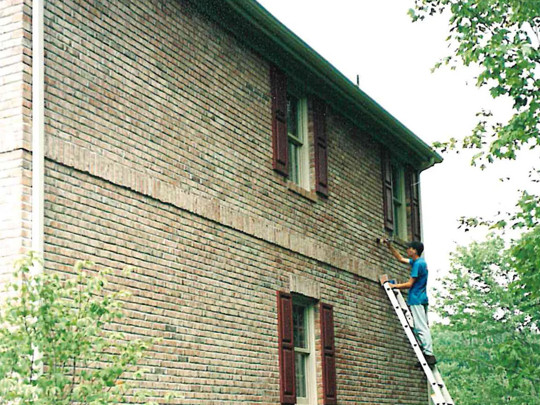 An applicator on a ladder working a brush across the tall side wall of the house in 1998, a band of darker brick running along the wall beside him