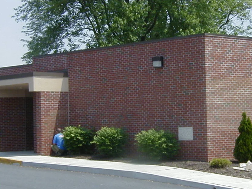 A single story entrance wing in 2004, its brick a dark variegated red with a stone plaque set in the wall, a canopied doorway at the left and shrubs along the base