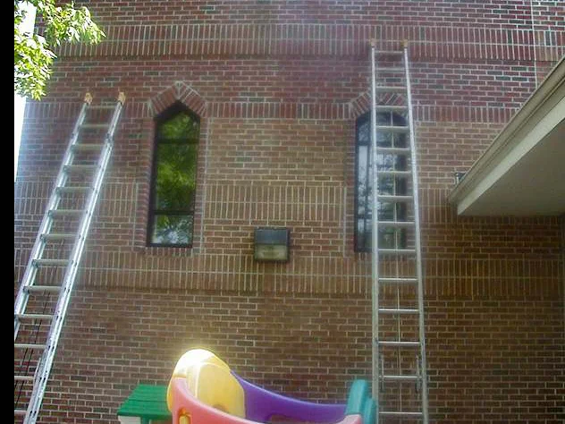 Two extension ladders standing against the second story wall either side of a pointed arch window in 2004, play equipment on the ground below