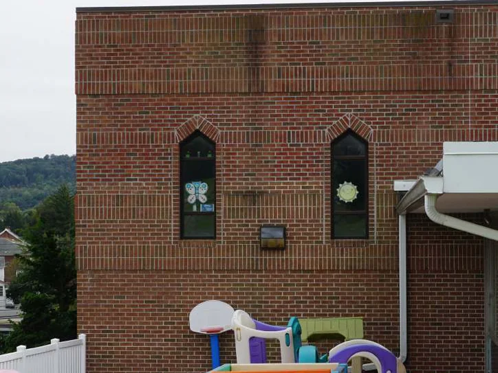 Two pointed arch windows on the second story in 2019 with paper decorations in the glass, the brick one even range, play equipment and a basketball hoop below