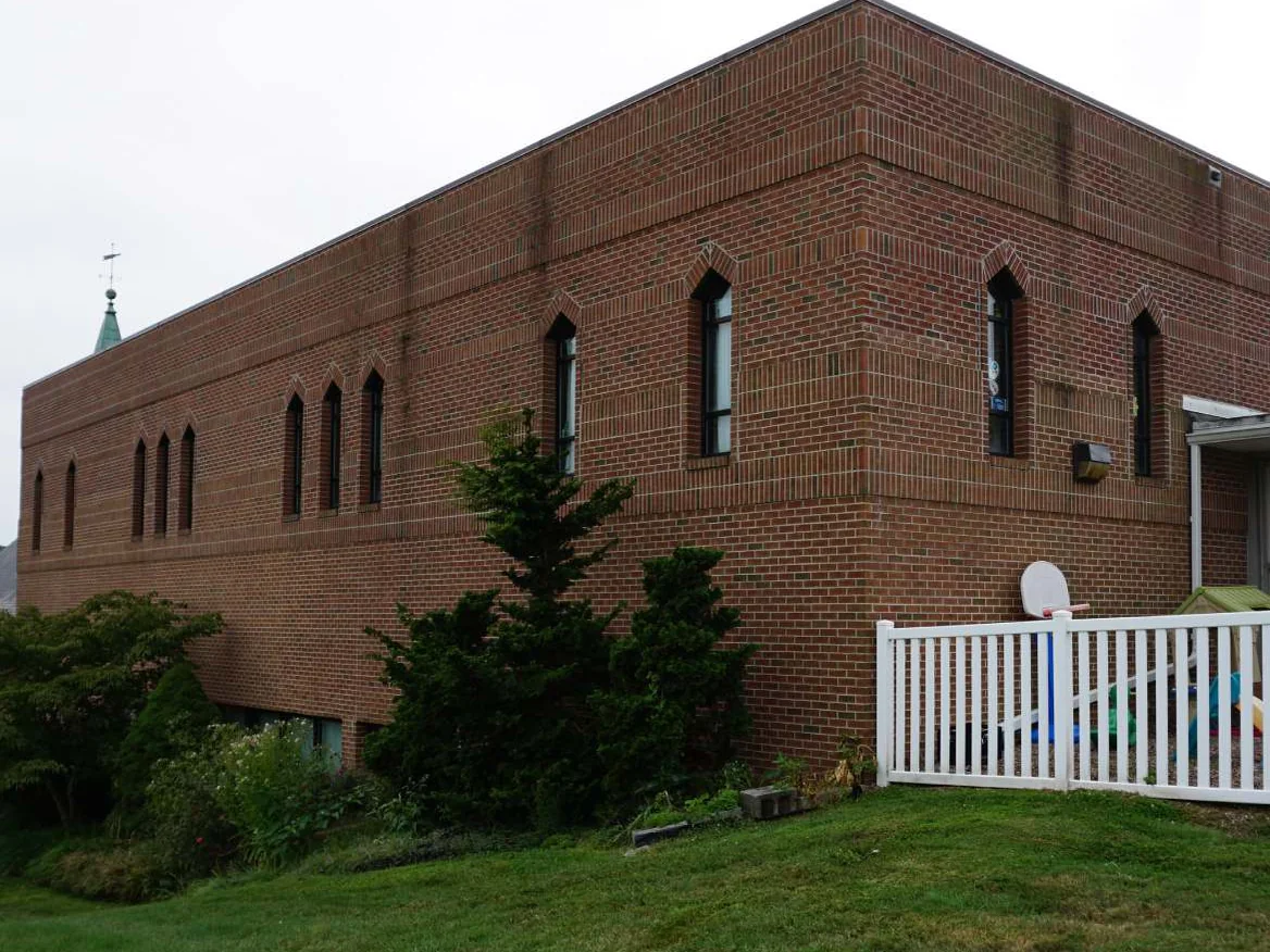 The long side of the church in 2019, two stories of red brick under a flat roof with pointed arch windows along the upper floor, a steeple behind and a white fenced play yard at the corner