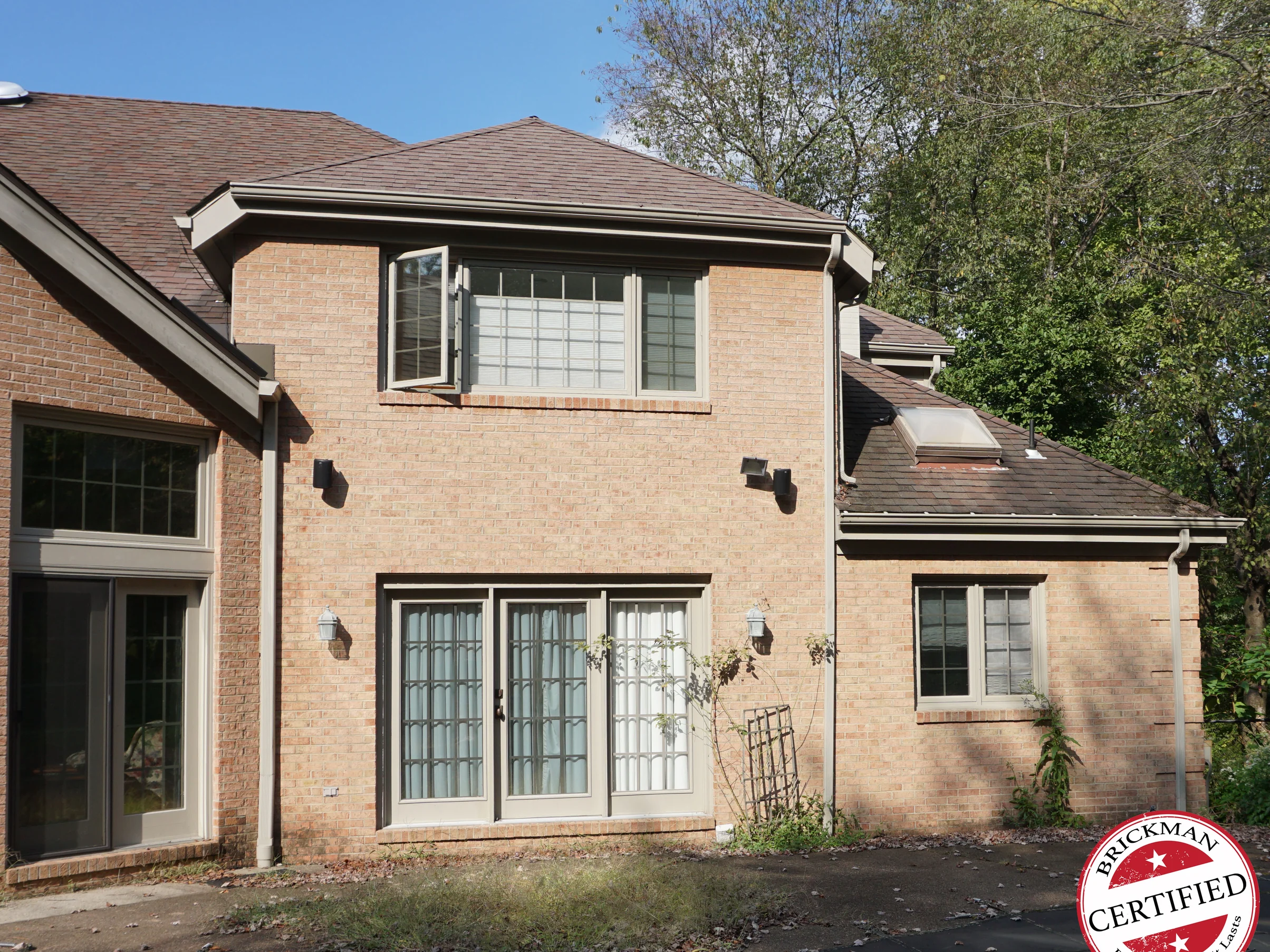 The back of the house in 2019, the two-story section with its french doors, the tall window at the left and the single-story wing at the right all reading as one even buff and tan range, with the certification stamp at the corner of the frame