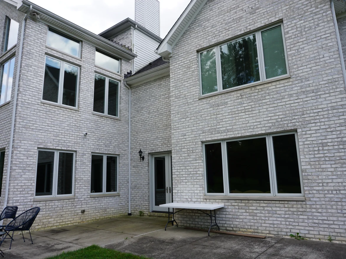 The rear of the house in 2019, two storeys of pale grey brick meeting at an inside corner above a concrete patio