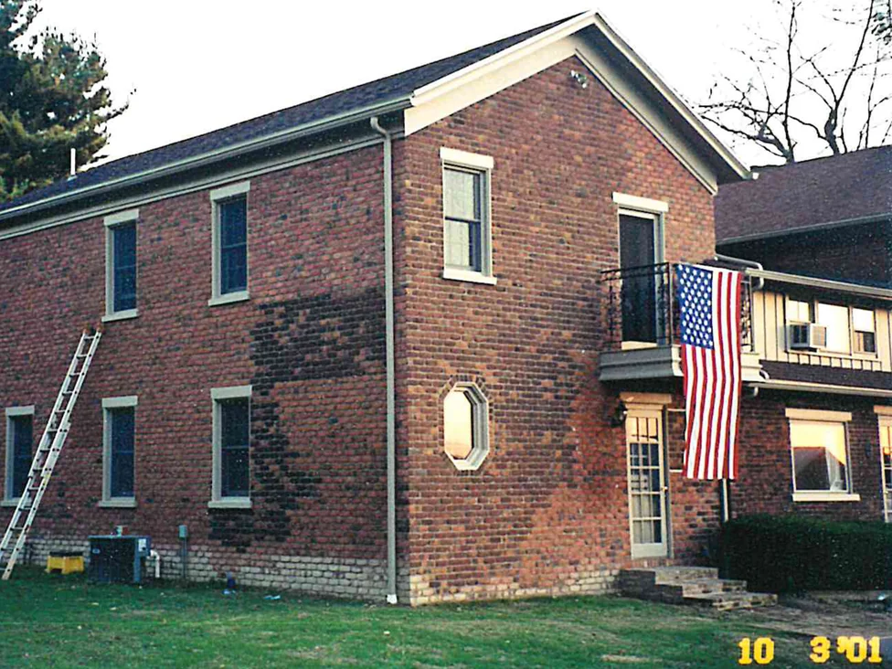 The gable end and long wall of a two-story brick house in 2001, with a tall block of much darker brick standing in the middle of the long wall and a ladder leaning at the far end