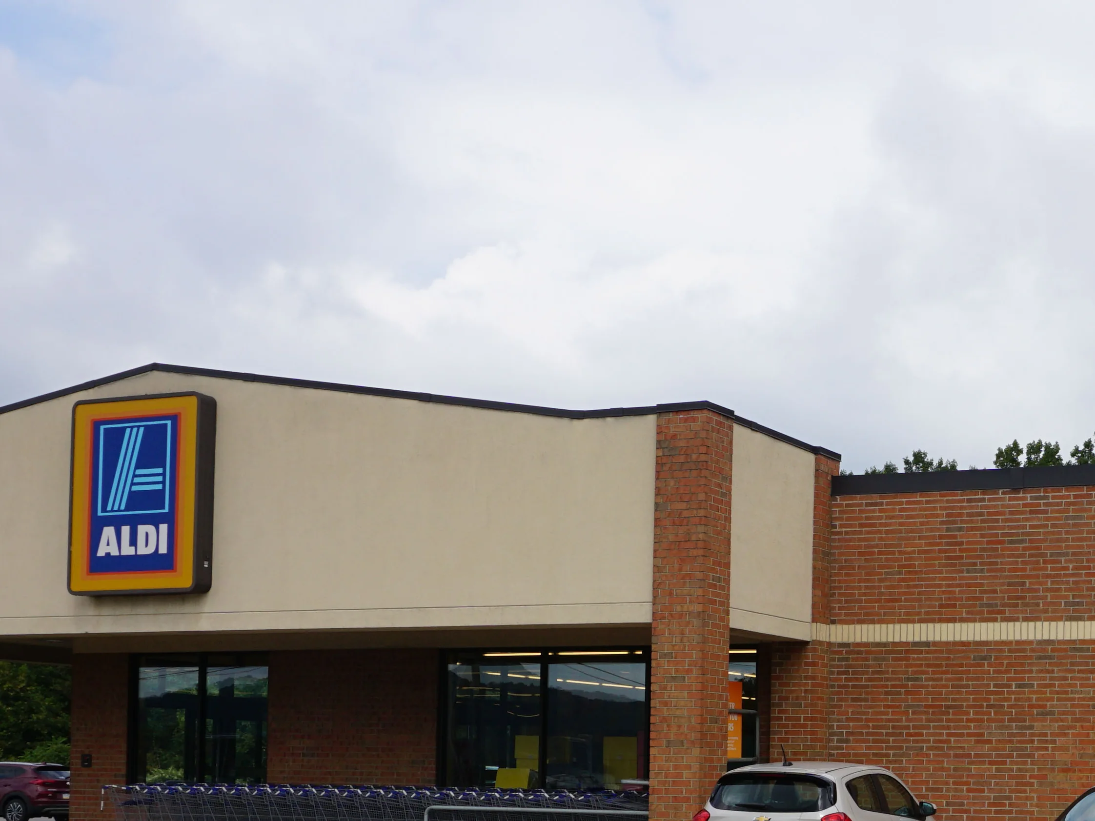 The front of the store in 2019, its lit ALDI sign over the entrance canopy, the carts racked along the glass and the brick pier and wall beside them