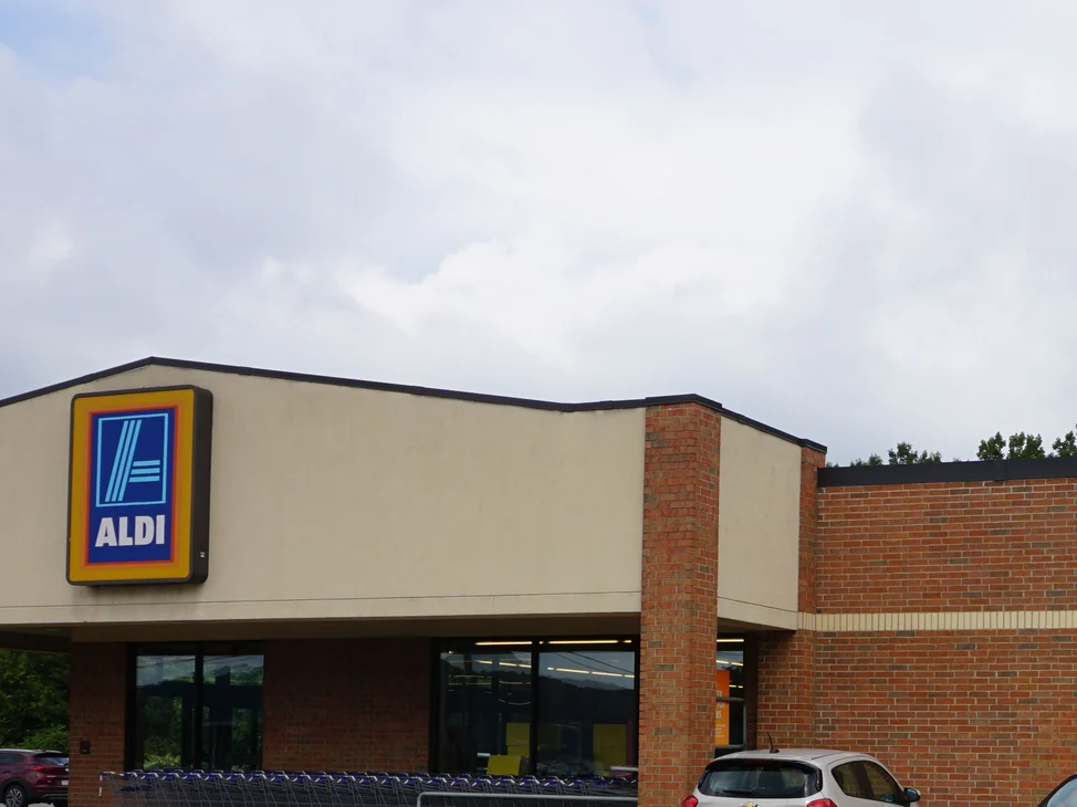 The front of the store in 2019, its lit ALDI sign over the entrance canopy, the carts racked along the glass and the brick pier and wall beside them.