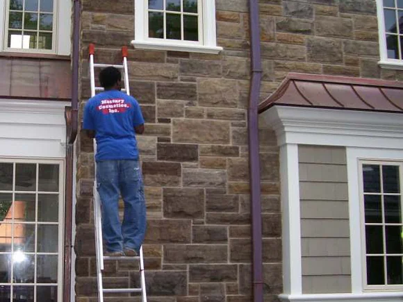 An applicator on an extension ladder in a Masonry Cosmetics shirt, working stain into the upper stone by hand