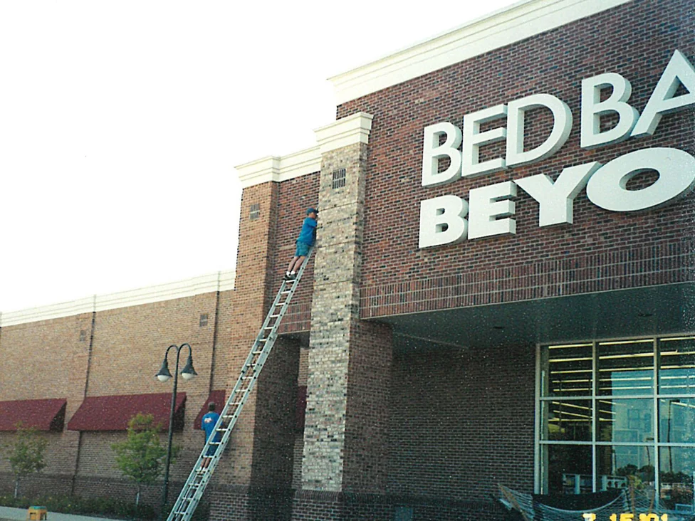 Two applicators on ladders at the store entrance in 2001, one pier already carried to a dark red brown beside a second still in its original cream