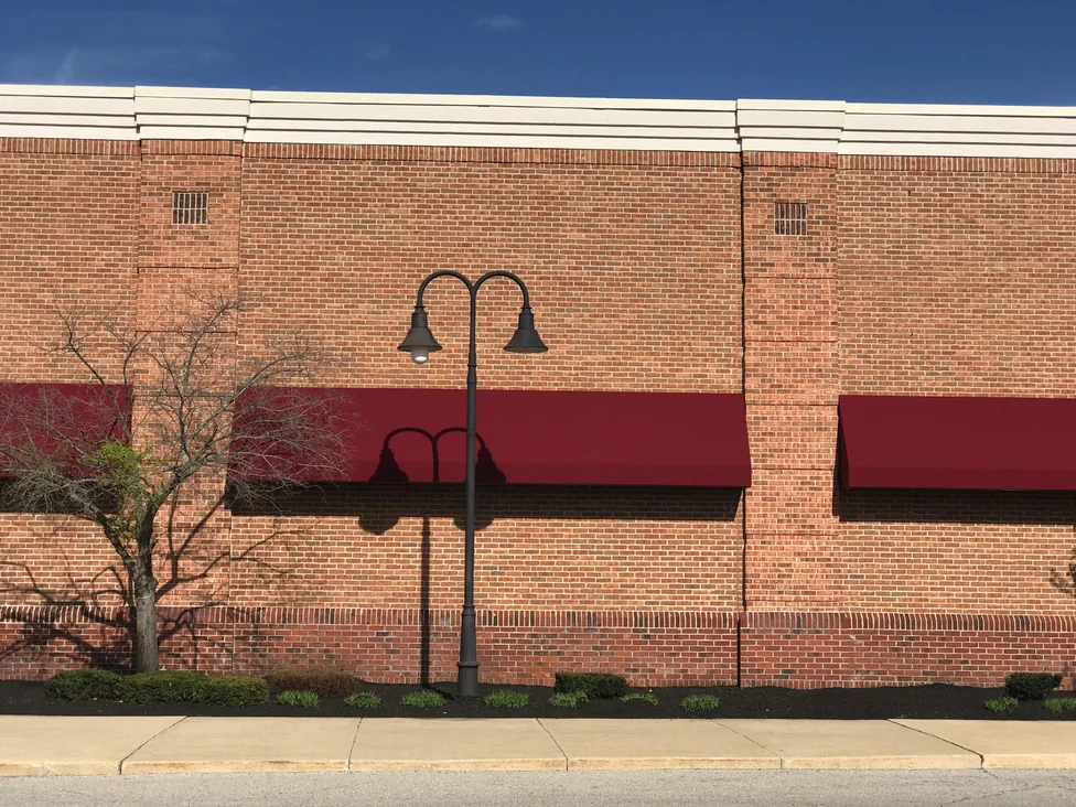 A bay of the same wall close up in 2019, a projecting brick pier between two maroon awnings with a lamp post standing in front of it
