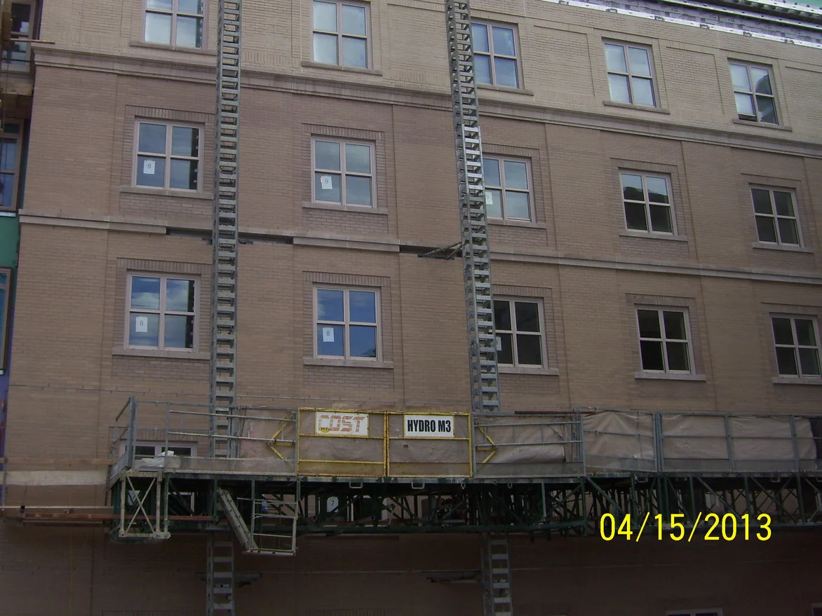 Three floors of the college building seen from a suspended swing stage in 2013, the brick a pink brown, with paper floor markers taped inside the windows