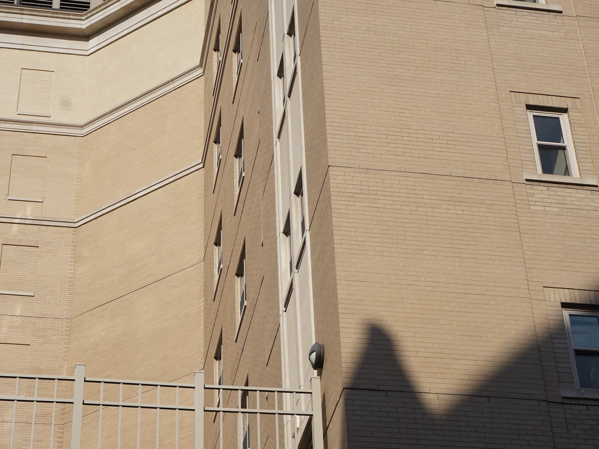 An outside corner of the college in 2019, a broad blank brick wall running up to a cast stone band, the brick an even warm tan across both faces