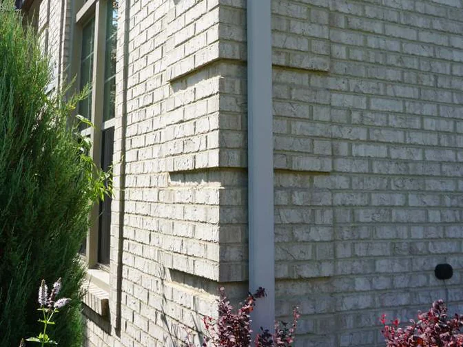 The same outside corner in 2019, one pale range of brick on both faces beside the downspout, barberry at the foot