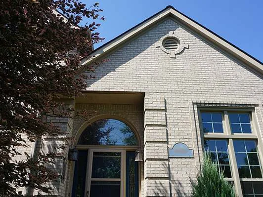 The entry gable of the house in 2019, pale grey brick around the arched entry, a purple-leafed tree in front