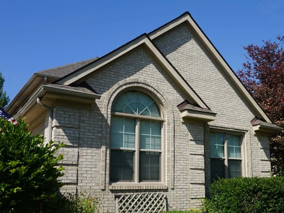 The front gable of the house in 2019, pale grey cut and rollback brick around an arched window with a lattice panel below it, shrubs and a stone-edged bed in front