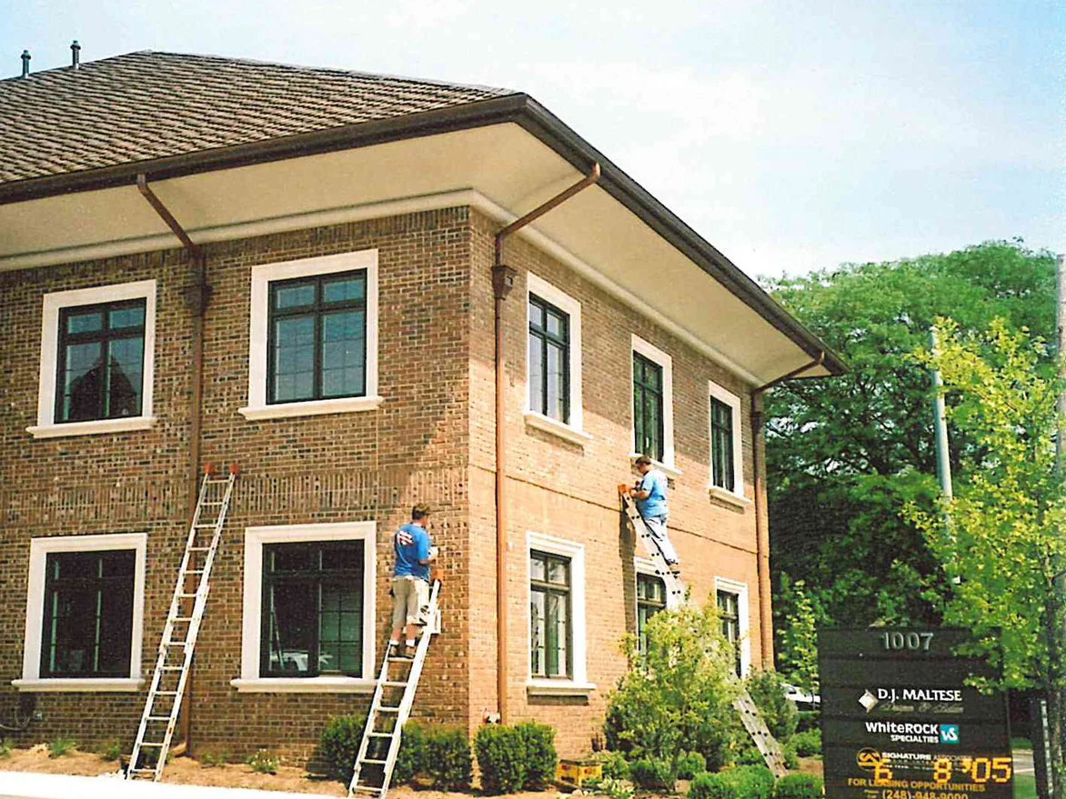 Two applicators on ladders working across the brick of a two-story office building, its brick a pale uneven tan