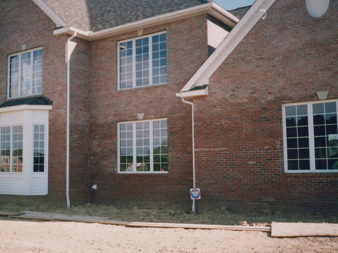 The bay window elevation in 1996, a white clad bay at the left, keystoned grid windows in a red brick wall with patchy pale streaking, boards laid on the dirt below