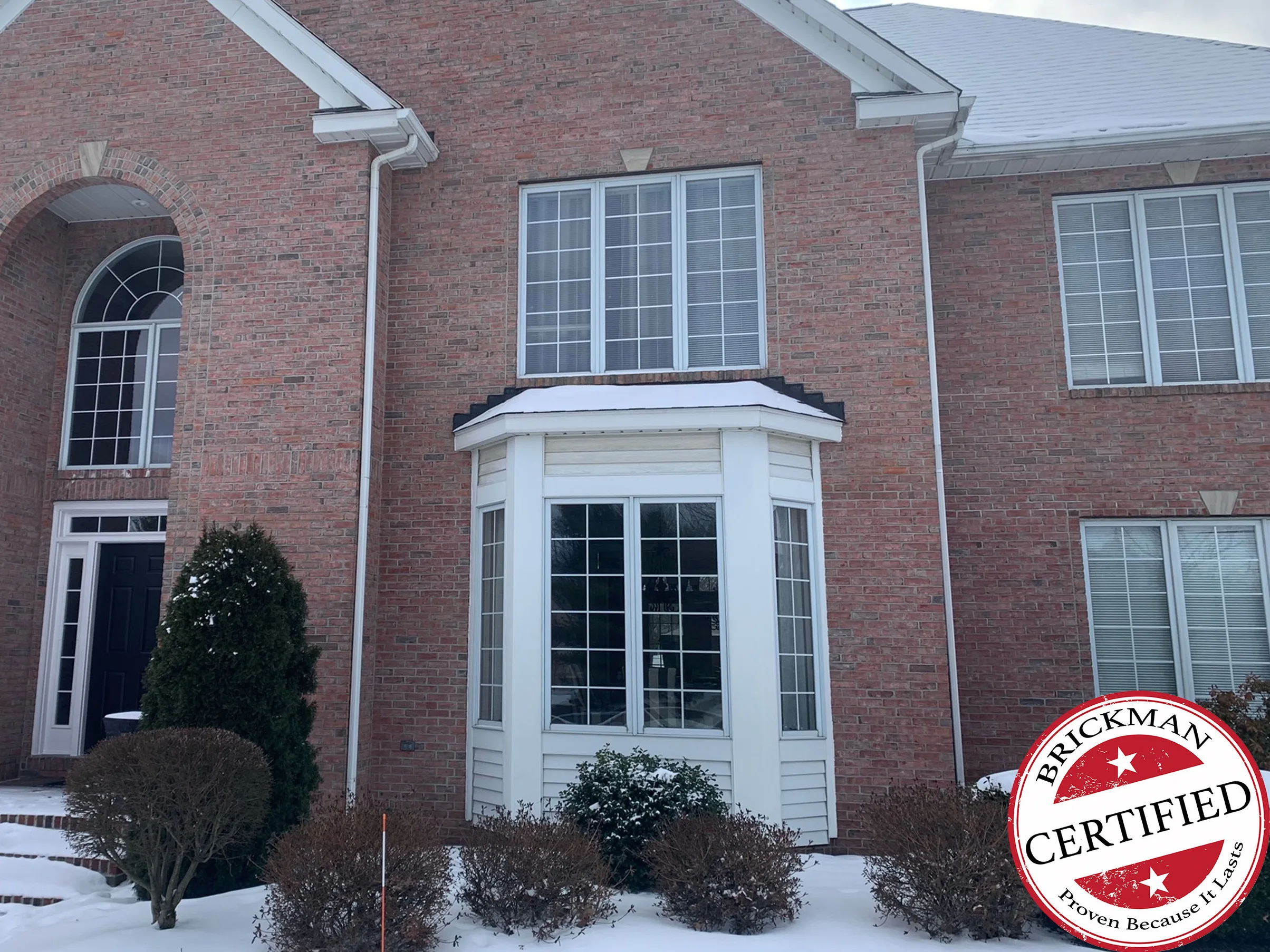 The front of the two-story brick house in 2021 with snow on the ground, a white clad bay window under a keystoned window beside the arched entry, and the certification stamp in the corner