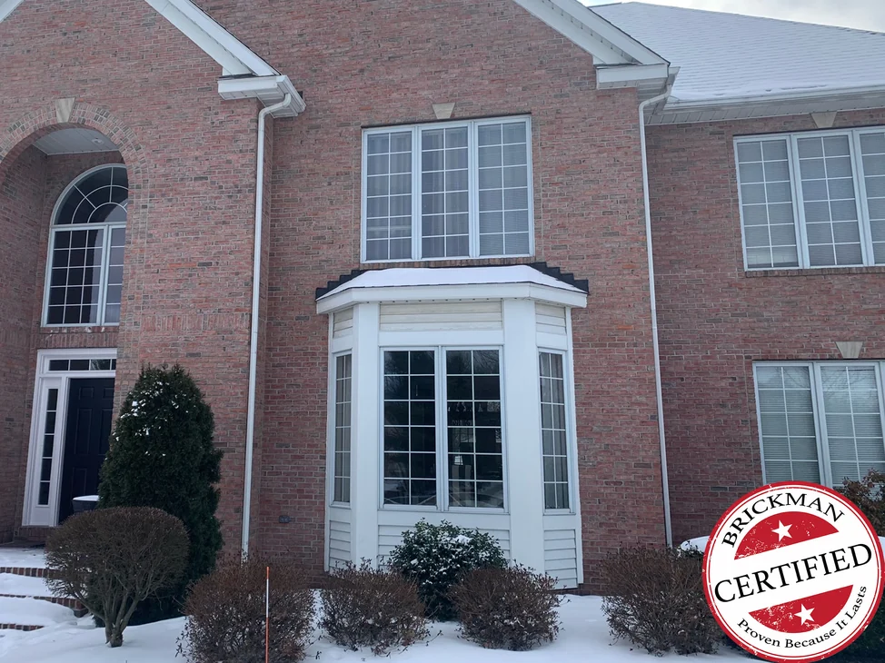 The front of the two-story brick house in 2021 with snow on the ground, a white clad bay window beside the arched entry, and the certification stamp in the corner