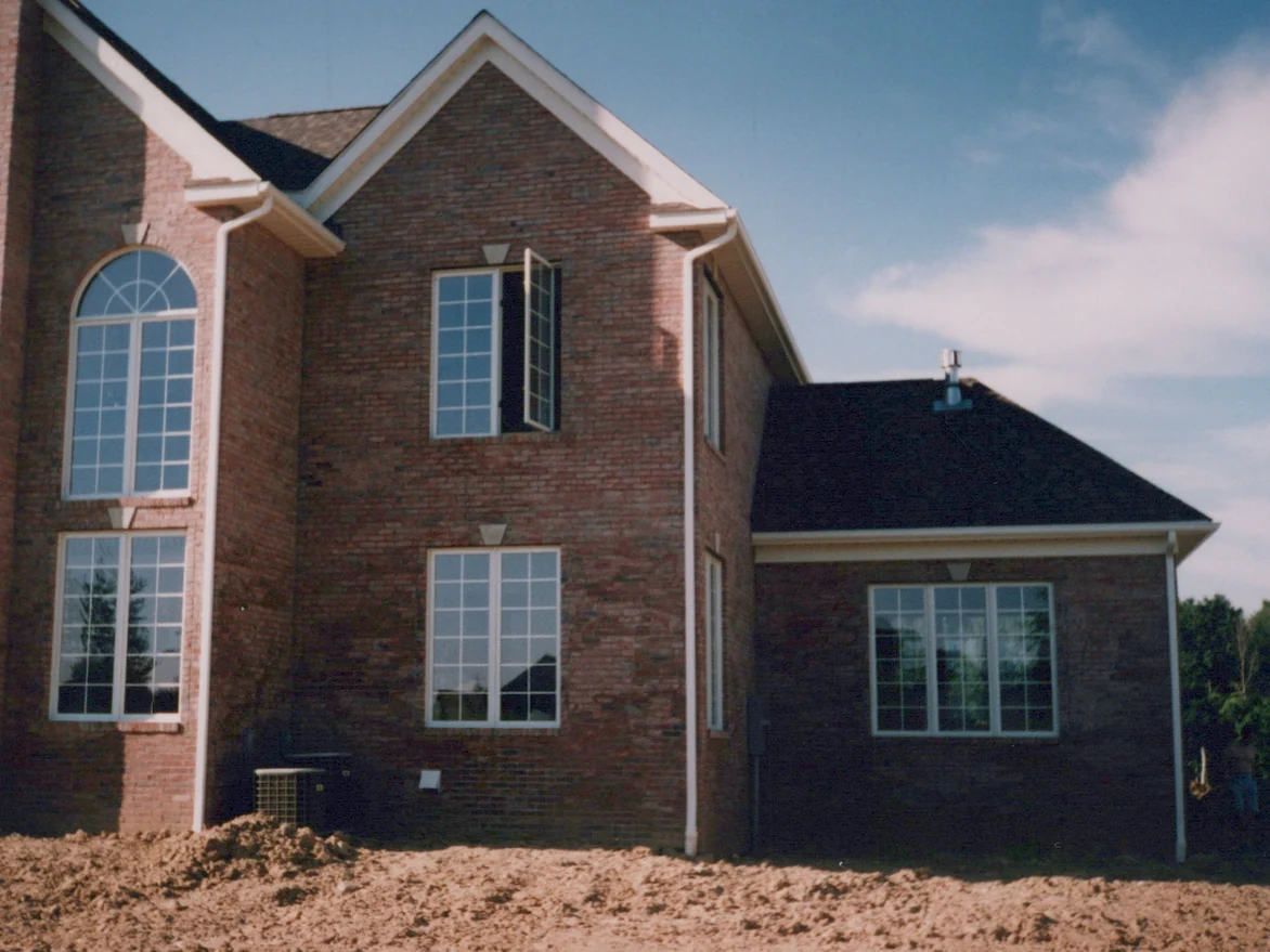 A gabled elevation of the new house in 1996 on bare graded soil, the red brick streaked and blotched pale across the face, an air conditioning condenser at the foot of the wall