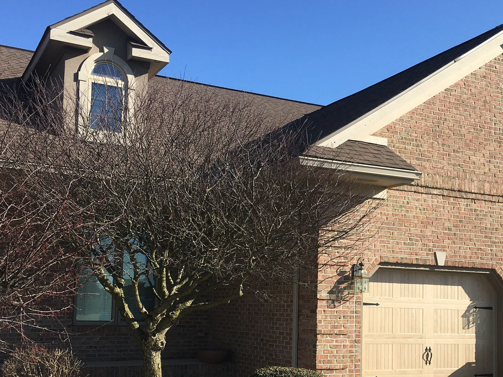 The garage gable of the brick house in 2021 under a clear sky, a bare tree in front of the picture window and a stucco dormer above the roofline