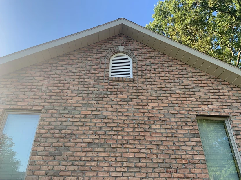 The peak of the same gable in 2019 seen from below, the arched louvre vent set in a soldier arch with the brick running one range up to the soffit against a clear sky