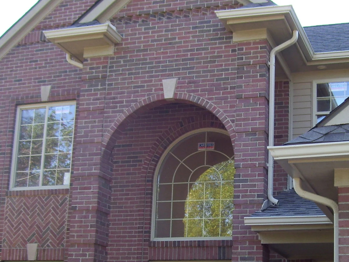 The gabled entry after staining, the arch, the pier and the wall beside them in one darker red range, cream corbels above and a herringbone brick panel under the left window