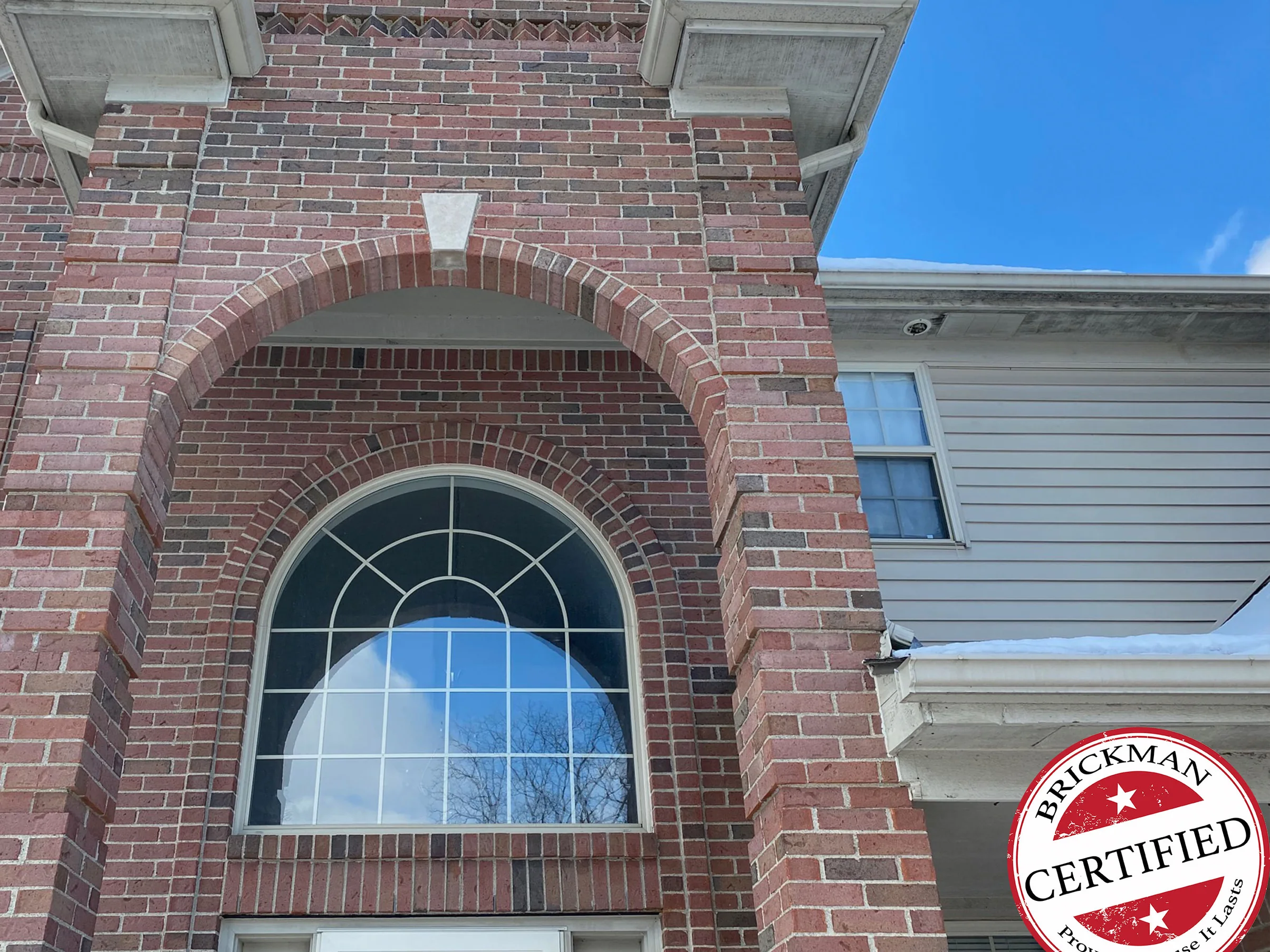The entry arch in 2021 with snow on the roof beyond, red brick from the gable down the pier in one range around the arched transom window, with the certification stamp in the corner