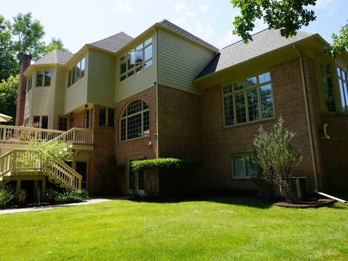 The rear of the house in 2019, a red brick lower level under cream siding with a raised deck and an open stair