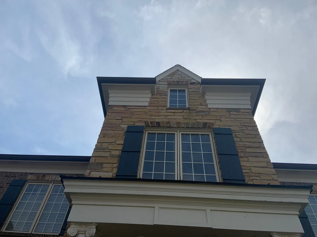 Looking up at the stone dormer and shuttered window above the entry portico in 2019, with brick and a shutter at the left edge