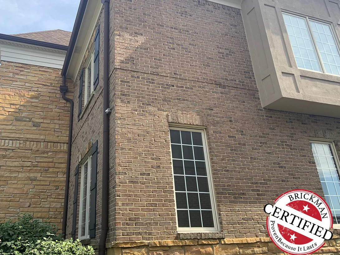 An outside brick corner of the house in 2019 beside the stone wing, the units an even tan and brown range with pale mortar joints, carrying the certification stamp