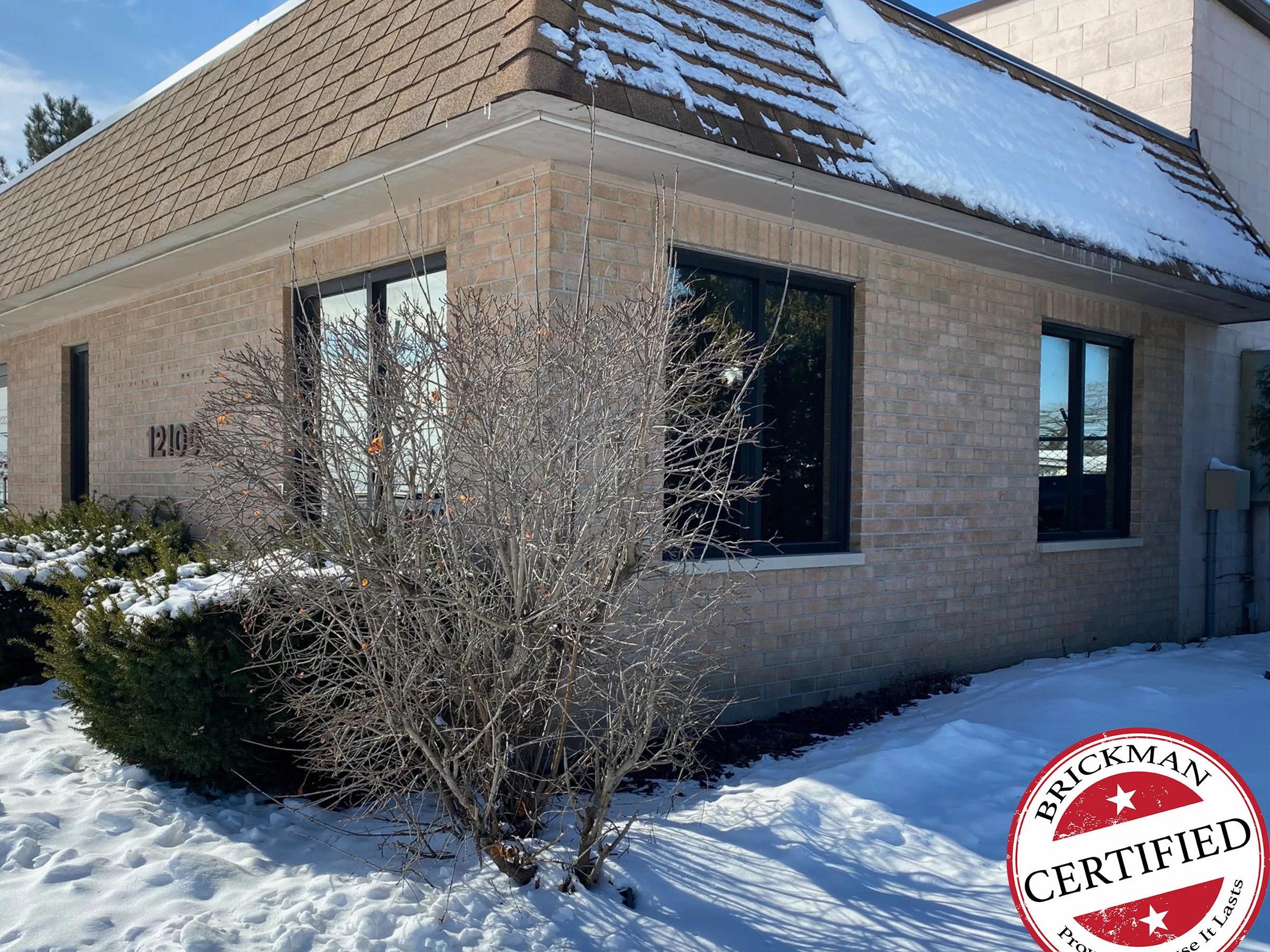 The corner of the building in 2021 with snow on the ground and the shrubs, buff brick reading one range across both elevations under a shingled hip roof, with the certification stamp in the corner