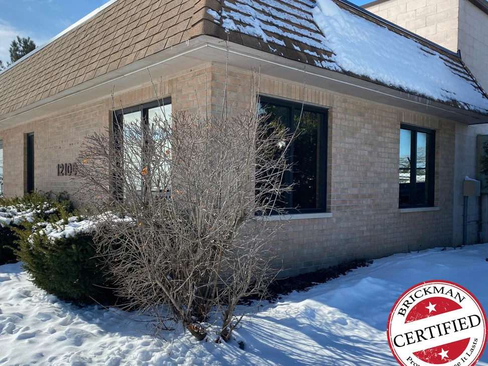 The corner of the Romulus building in 2021 with snow on the ground and the shrubs, buff brick reading one range across both elevations under a shingled hip roof, with the certification stamp in the corner