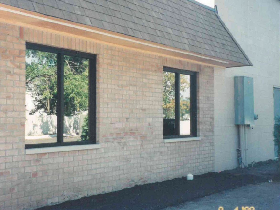A side elevation of the building in September 1999, buff brick between two dark framed windows under a shingled roof, with darker marks on the wall below the second window and a bare soil bed at the base