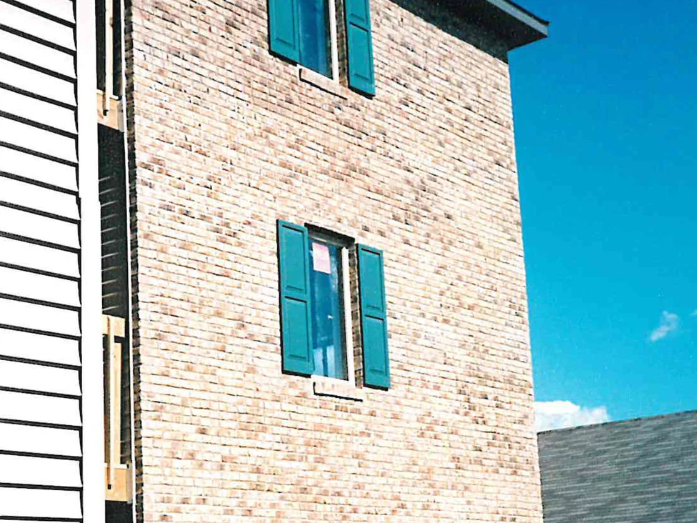 The brick end wall of an apartment building in 1998, the buff units running patchy and uneven between three stacked windows with teal shutters