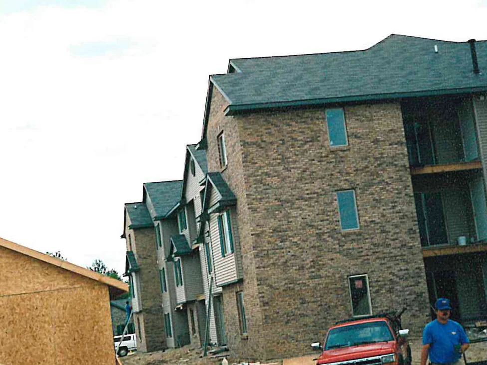 The apartment complex under construction in 1998, one finished brick end wall beside a run of dormers and siding, with a pickup and bare ground in front