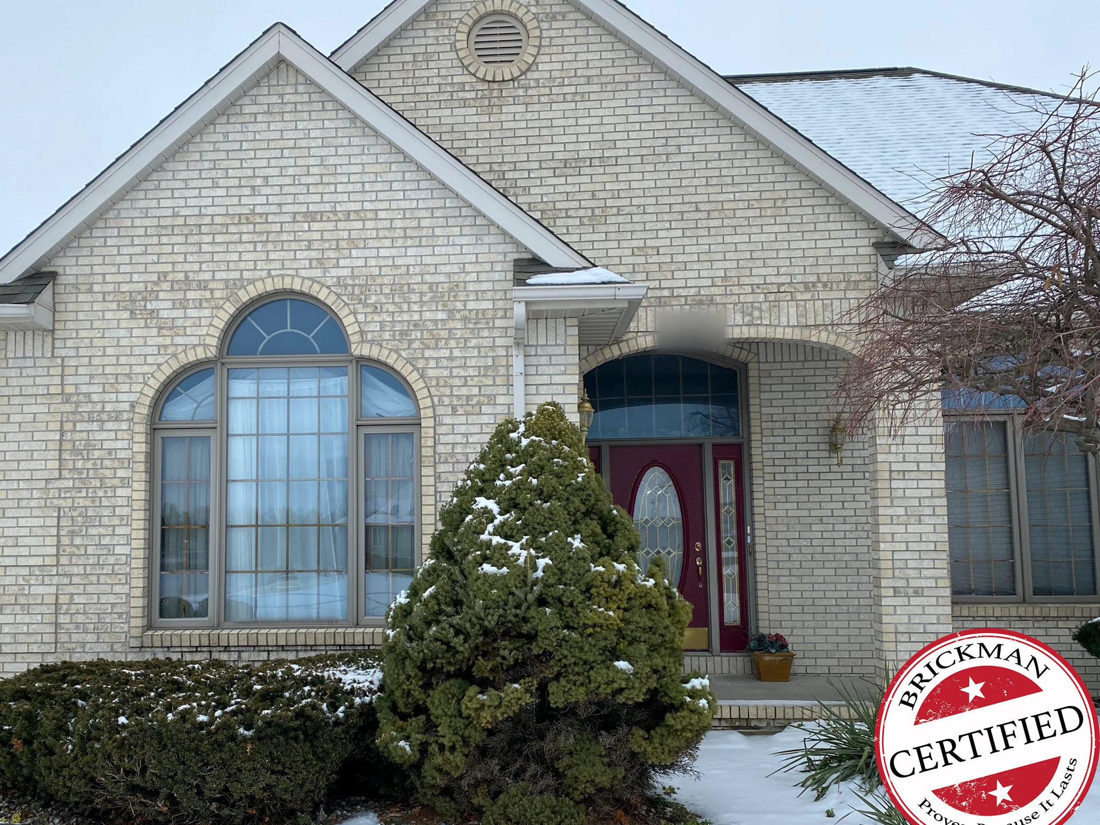 The front gable and the entry in 2020 with snow on the shrubs, the brick one pale grey and cream range from the arched window to the door, with the certification stamp in the corner