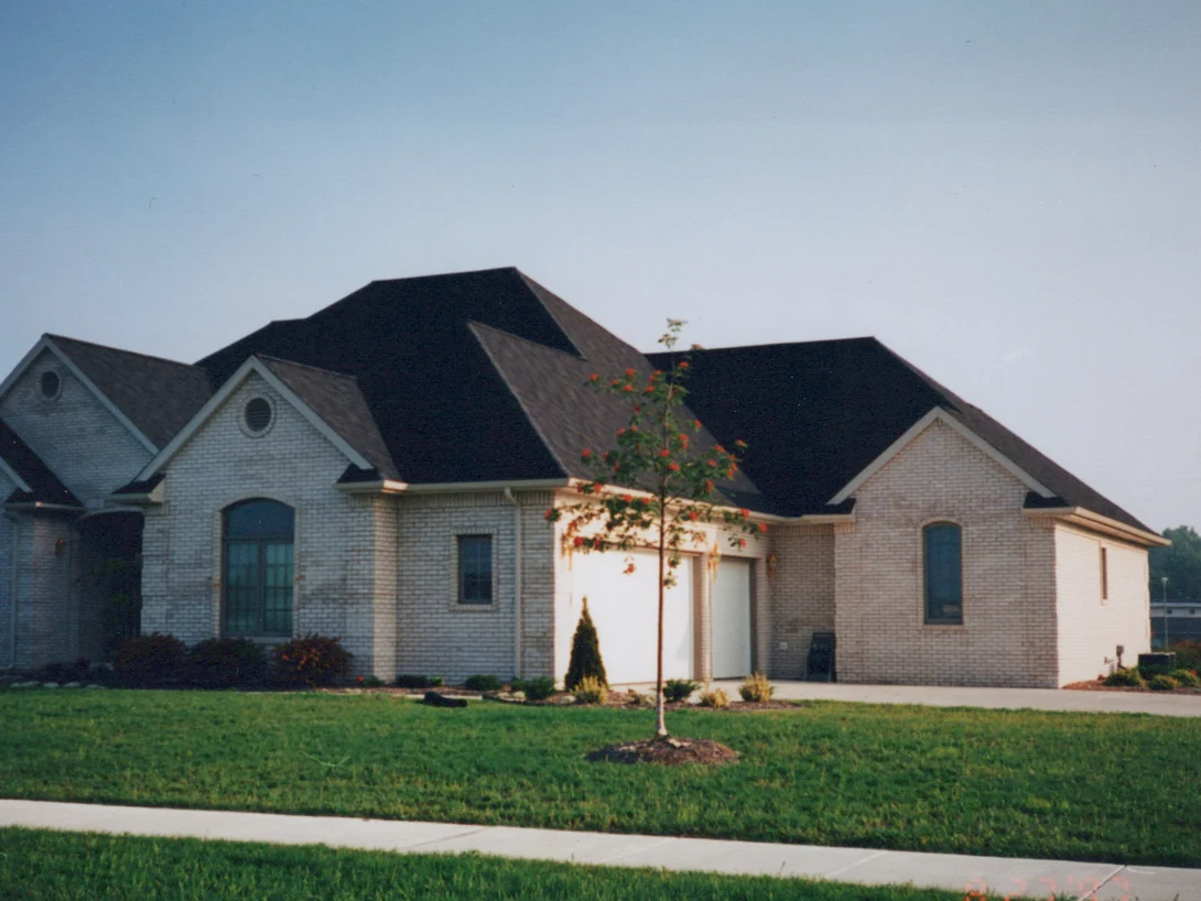 The whole front of the house from the street in 1997, three brick gables and a two-car garage under a dark roof, the brick a warm tan with a young tree on the lawn