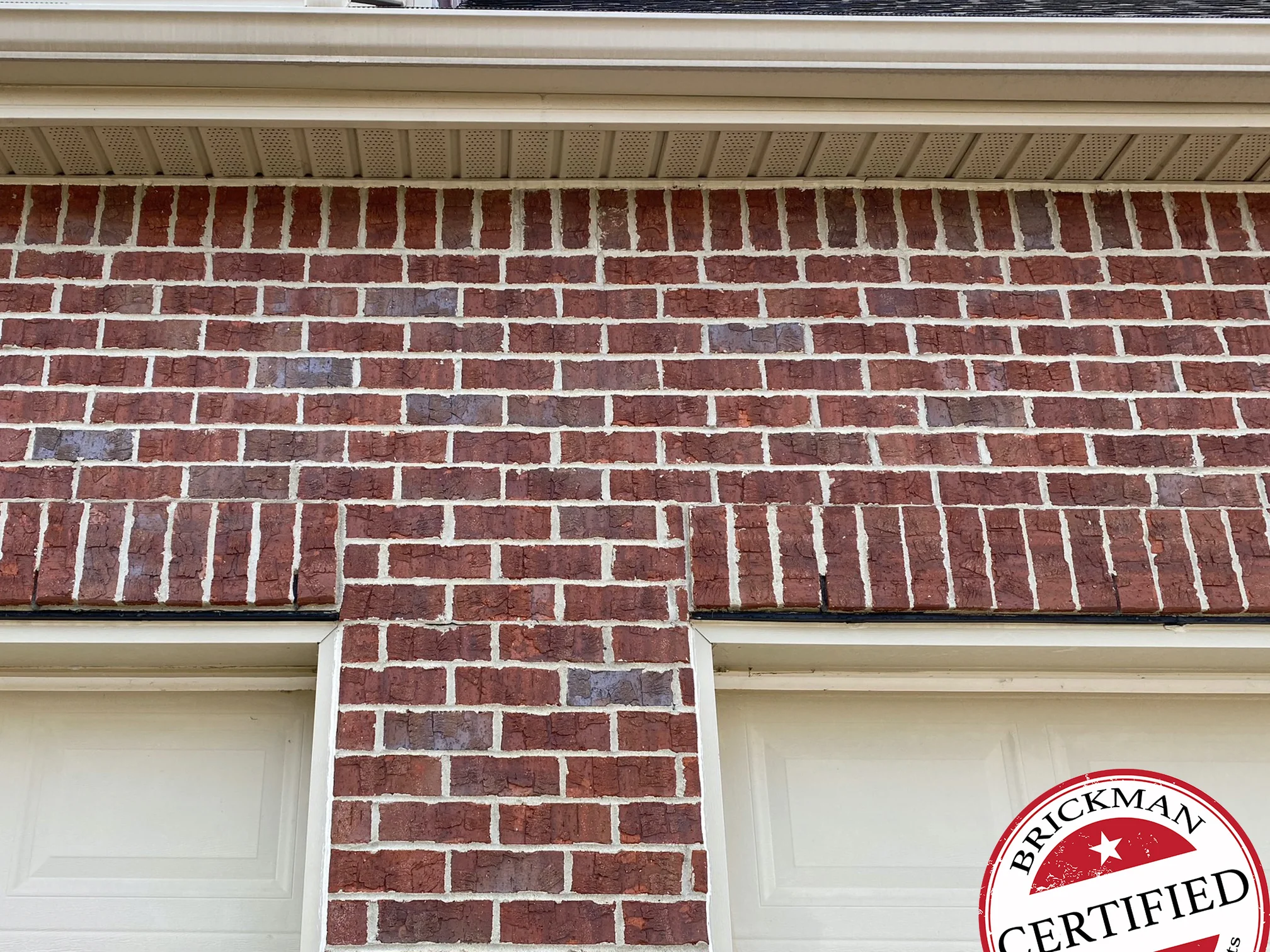 The brick over the two garage doors in 2020, one red range across the courses under the soffit and down the pier between the doors, with the certification stamp in the corner