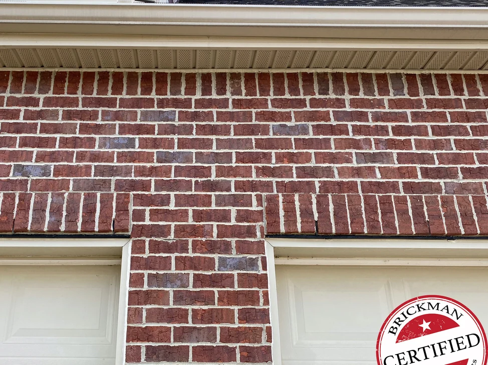 The brick over the two garage doors in 2020, one red range across the courses under the soffit and down the pier between the doors, with the certification stamp in the corner.