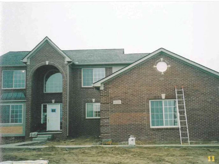 The whole front of the two-story house in 2003, arched entry and gabled wing complete, a ladder standing against the right gable and bare graded dirt across the yard