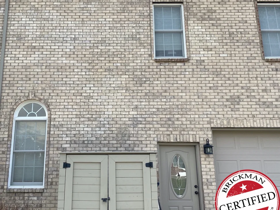 The garage wall of the Saline house in 2020, buff brick one range from the arched window past the entry door to the garage, with the certification stamp in the corner.