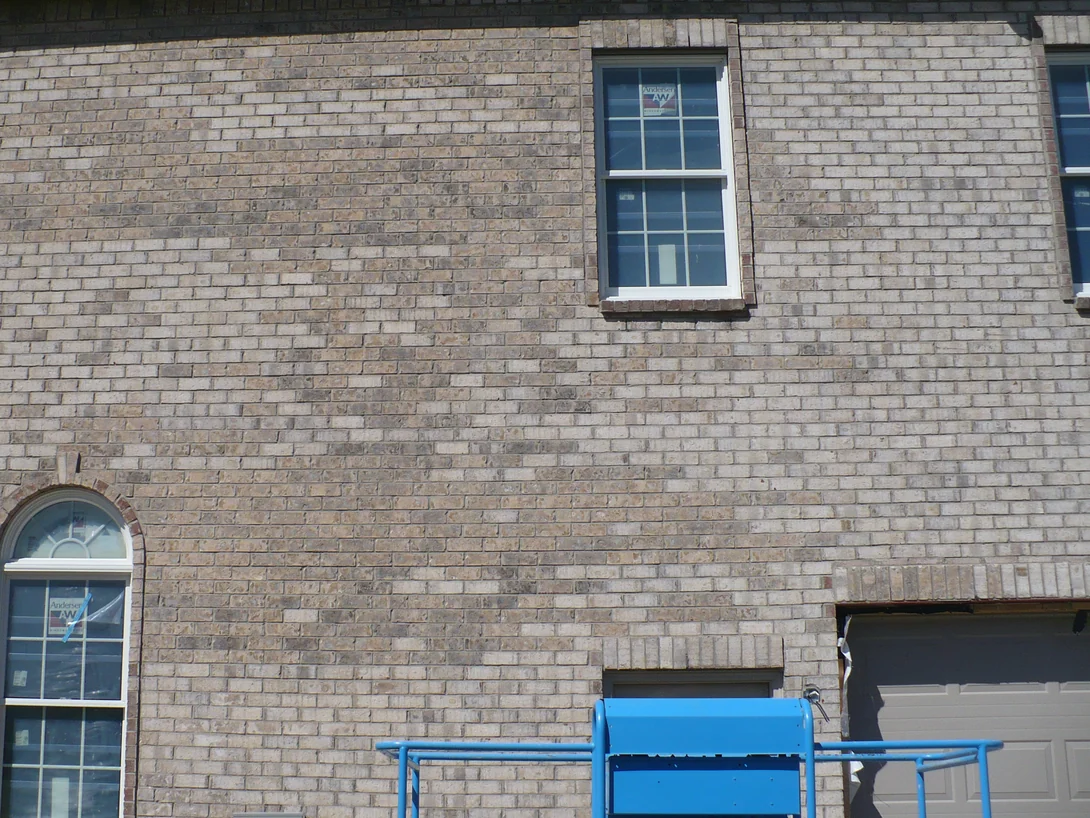 The garage wall of the house in 2012 before staining, buff brick with clusters of much darker units across the upper wall and a blue lift parked below the window