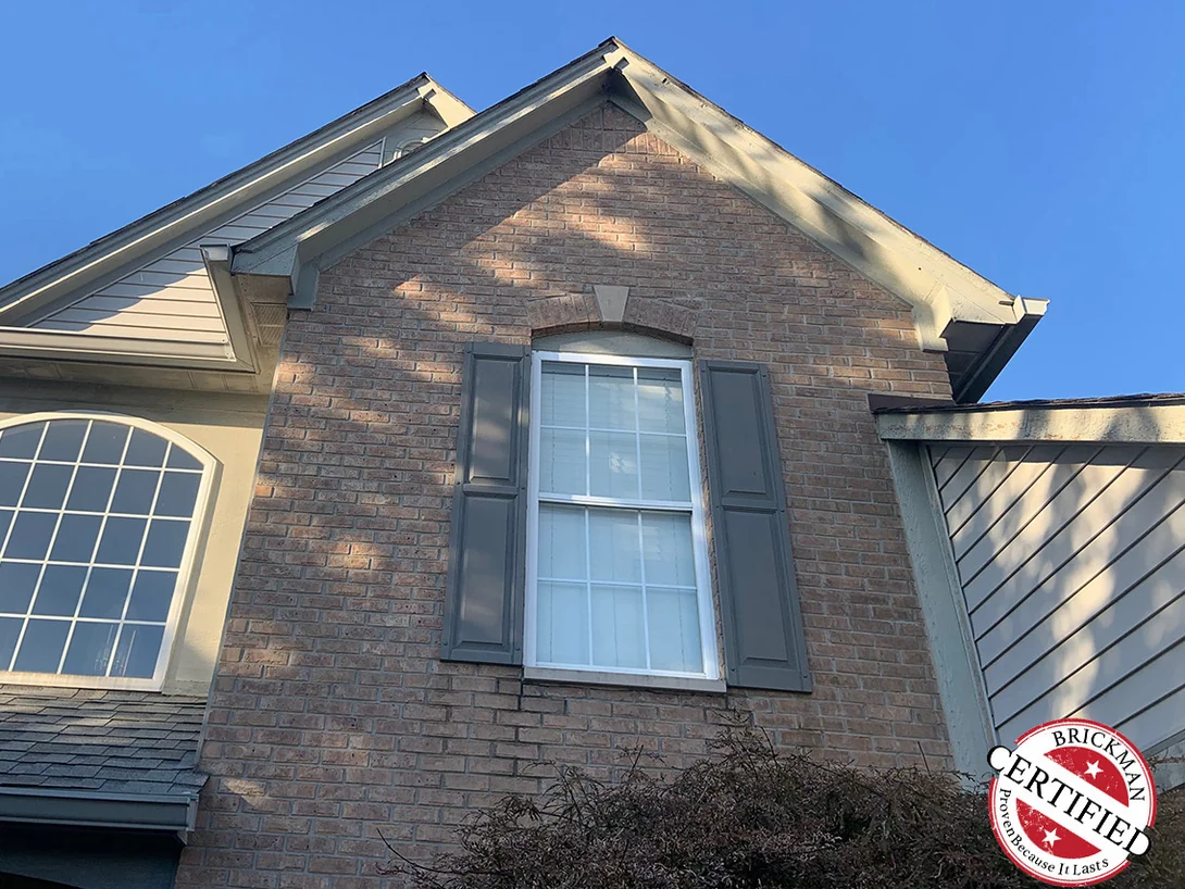 A front gable in 2019 with an arched brick head over a shuttered window and a tall arched window on the bay beside it, the certification stamp at the corner