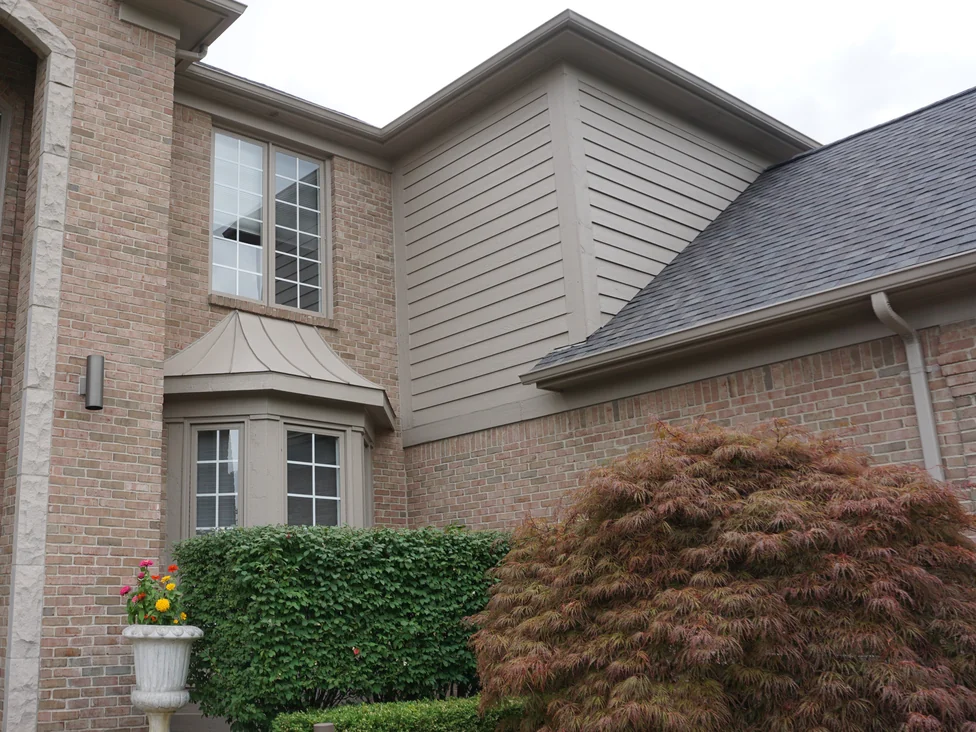The bay window under its copper roof in 2019, the brick around it and along the wing to the right holding one tan and pink range above a clipped hedge