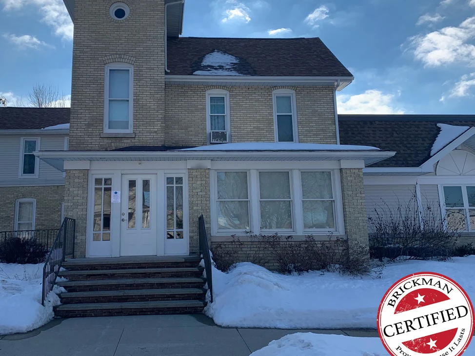 The front of the Shell Rock house in 2021 under snow, one cream brick range across the tower, the two-story wall and the entry, with the certification stamp in the corner.