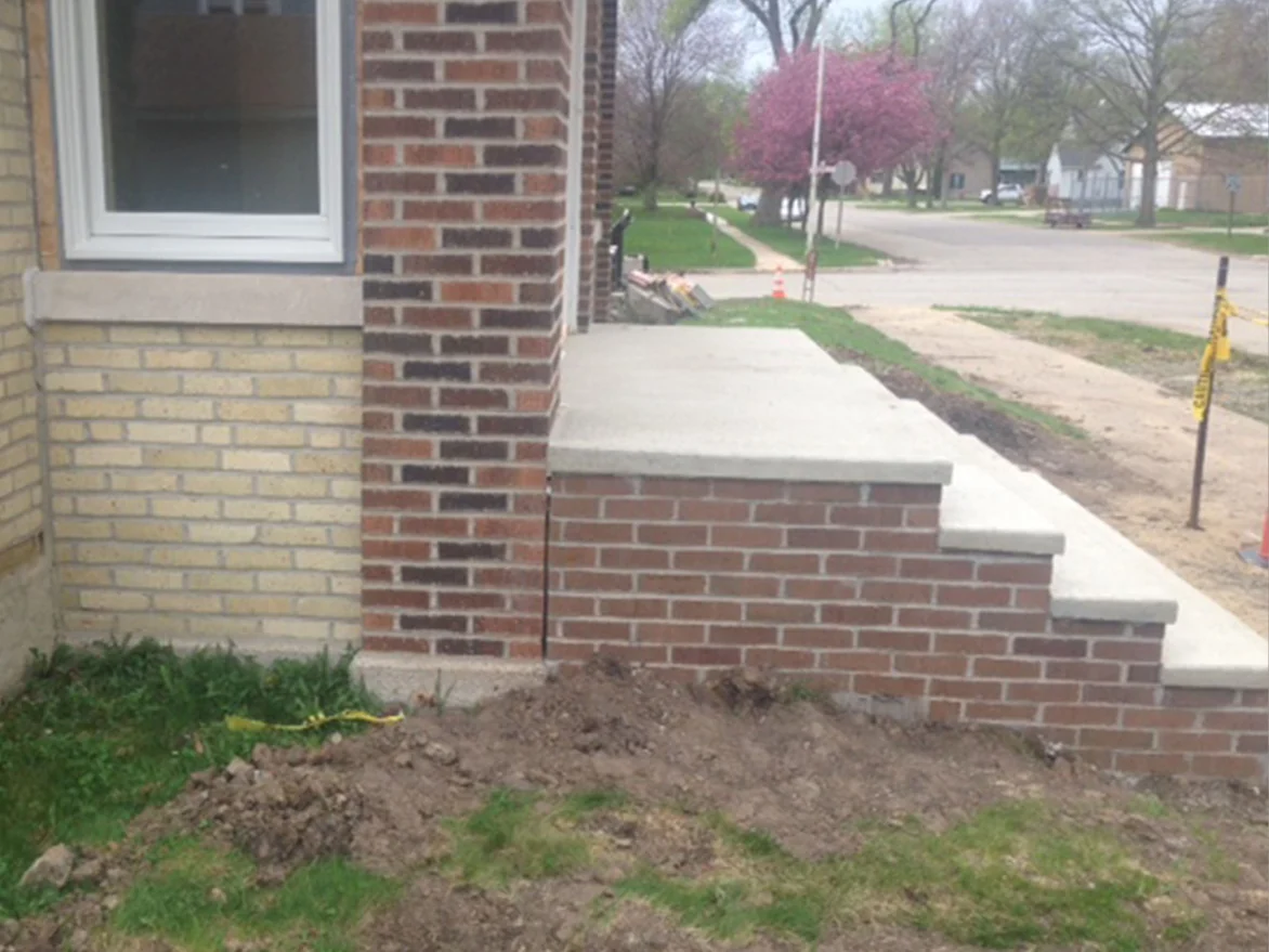 A red brown brick pier and porch wall built against the cream brick of the house in 2013, new concrete steps beside them and bare dirt in front