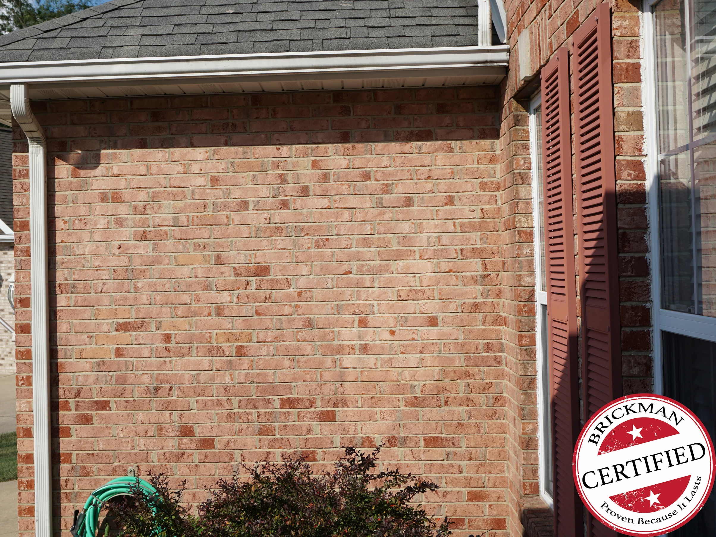 The side wall of the house in 2019, its brick one even light red and tan range from the ground to the eaves, with a window and shutters past the outside corner