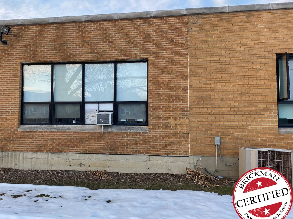 The window bay in 2021 with snow on the ground, tan brick running from the stained section across the vertical joint into the existing wall, with the certification stamp in the corner